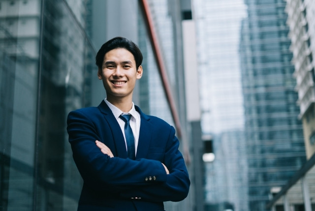 スーツを着た若いビジネスマンがオフィス街で自信を持って微笑む姿。外国人正社員の採用成功やキャリア形成、企業での活躍をイメージさせる場面