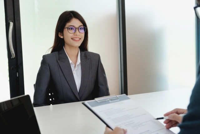 スーツ姿の若い外国人女性が就職面接で笑顔を見せる場面。面接官が履歴書を確認しながら対話しており、企業の外国人採用や人材活用をイメージした写真