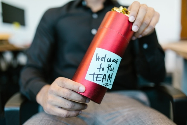 入社初日に新しい外国人正社員を歓迎する場面。赤いギフトボックスに“Welcome to the TEAM”と書かれたメッセージが添えられ、企業のオンボーディングや採用後の定着支援を表現