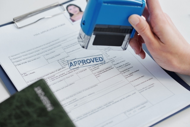 ビザ申請書に「APPROVED（承認済み）」のスタンプを押す手元。就労ビザや在留資格の許可を示すイメージ。