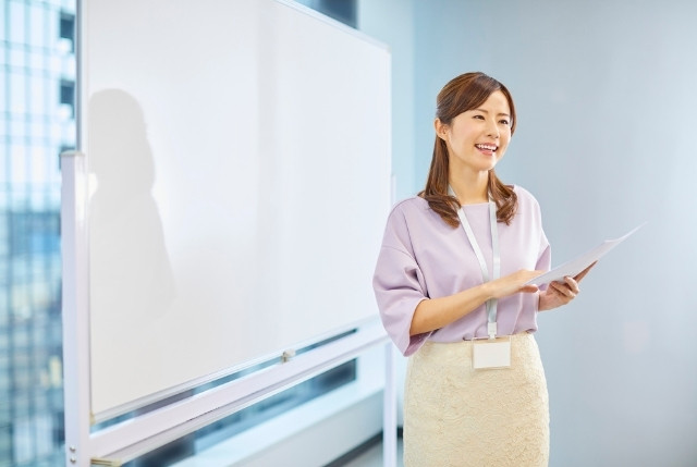 ホワイトボードの前で資料を手に笑顔で話す女性講師。生活オリエンテーションで外国人に説明する場面。